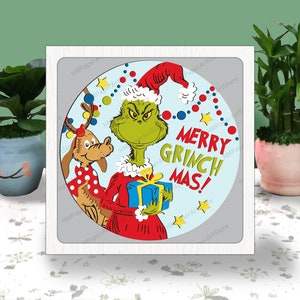 May include: A square white-framed wall art piece featuring a circular illustration of a green character in a red Santa suit and hat holding a blue gift box, next to a brown dog with antlers. The text reads MERRY GRINCH MAS in red and green.