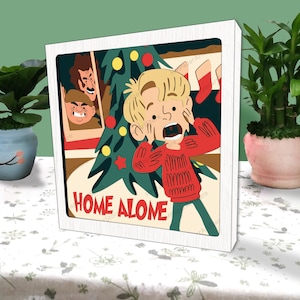 May include: A white framed illustration of a boy with blonde hair wearing a red sweater and looking scared. The boy is standing in front of a Christmas tree with the words "Home Alone" written below.