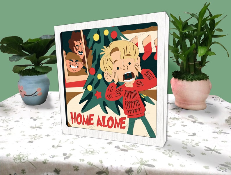 May include: A white framed illustration of a boy with blonde hair wearing a red sweater and looking scared. The boy is standing in front of a Christmas tree with the words "Home Alone" written below.
