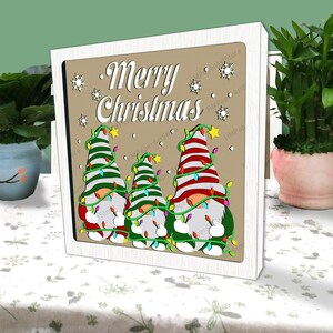 以下が含まれることがあります： 「メリークリスマス」と書かれた木製の看板と、赤と緑の帽子をかぶってクリスマスツリーを持った光るライトを持った3人のノームの楽しいシーン。ノームは雪片が降る茶色の背景に立っています。