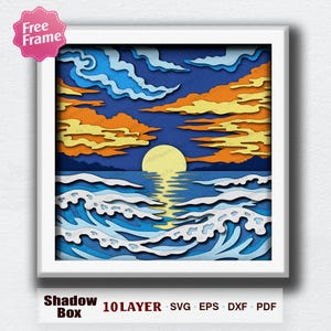 May include: A framed, multi-layered paper art piece depicting a sunset over the ocean. The artwork features blue, orange, and yellow hues, with stylized waves and clouds. The frame is white, and the piece is labeled "Shadow Box" with "10 LAYER SVG EPS DXF PDF".