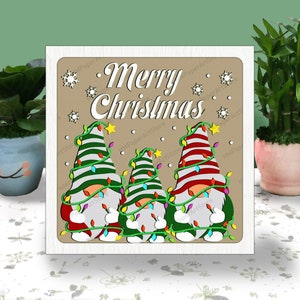 以下が含まれることがあります： 「メリークリスマス」と書かれた木製の看板と、赤と白のストライプの帽子、緑と白のストライプのシャツを着た3人のノーム。ノームは緑のライトがついた木を持っている。