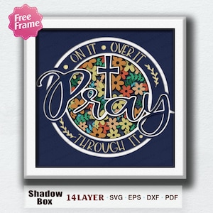 May include: A framed shadow box art piece with the word "Pray" in a cursive font, centered over a floral design and a cross. The text "On It, Over It, Through It" surrounds the floral design. The frame is white, and the background is navy blue.