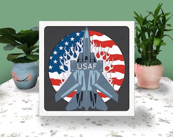 May include: A gray and blue fighter jet with the text "USAF" on the side, flying over a red, white, and blue American flag with flames.