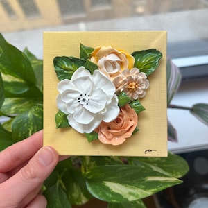 May include: A yellow canvas painting with a 3D floral design. The flowers are white, peach, and yellow with green leaves. The painting is signed "Jeni" in the bottom right corner.