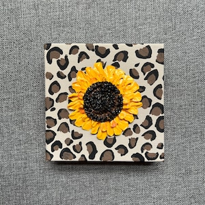 May include: A small canvas painting with a yellow sunflower with a black center on a brown and white leopard print background.