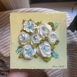May include: A hand-painted canvas with a textured background in a pale yellow color. The canvas features a bouquet of white roses with blue accents and green leaves. The artist's signature, "Olivia Elliot", is in the bottom right corner.