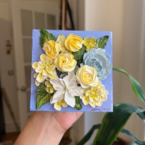 May include: A blue canvas painting with a floral arrangement of yellow and white flowers and blue flowers. The flowers are made of buttercream frosting.