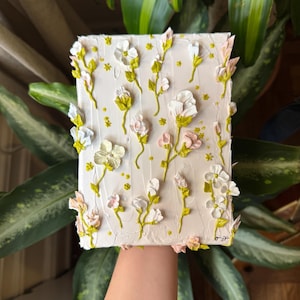 May include: A white canvas painting with a textured, raised floral design. The flowers are white, pink, and light blue with green stems and leaves. The background is a light cream color. The painting is signed "0-22".
