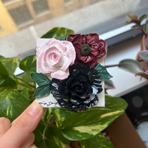 May include: A close-up of a handmade floral art piece featuring three flowers. The flowers are made of polymer clay and painted in shades of pink, black, and burgundy. The flowers are arranged on a white background.