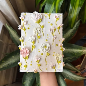 May include: A white canvas painting with a textured, 3D effect depicting a floral design. The painting features white and pink flowers with green stems and leaves.
