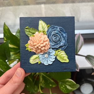 May include: A small canvas painting featuring a blue, pink, and white floral arrangement. The flowers are painted in a 3D style, giving the appearance of being sculpted.
