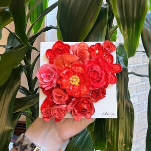 May include: A close-up of a red, white, and pink floral painting on a white canvas. The painting is made with thick, textured paint and features a variety of flowers, including roses, peonies, and carnations.