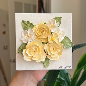 May include: A white canvas painting with a bouquet of yellow and white flowers. The flowers are painted in a realistic style and have a 3D effect. The painting is signed in the bottom right corner.