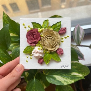 May include: A small canvas painting featuring a bouquet of flowers in shades of pink, white, and green. The flowers are painted in a realistic style and appear to be three-dimensional.