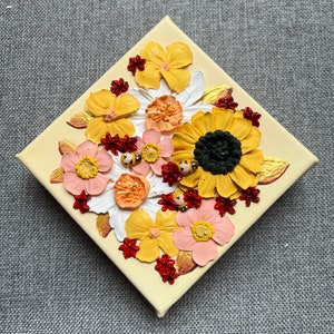 May include: A small canvas painting featuring a textured, 3D floral design. The painting depicts a variety of flowers in shades of yellow, pink, white, and red, with small black and yellow bees scattered throughout. The background is a light yellow.
