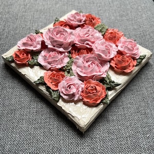 May include: A close-up of a small canvas painting featuring a cluster of pink and coral roses with green leaves. The roses are painted in a 3D style, giving the painting a textured look.