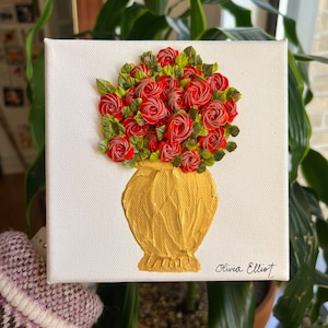 May include: A painting of a bouquet of red roses in a gold vase. The painting is on a white canvas and is signed "Olivia Elliot".