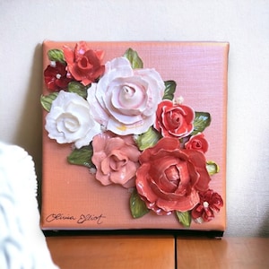 May include: A square canvas painting with a pink background and a 3D floral design. The flowers are made of white, pink, and red clay and are arranged in a cluster. The artist's signature, "Olivia Elliot", is in the bottom left corner.