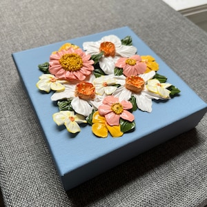May include: A square, light blue canvas features a 3D floral arrangement. The flowers are in shades of white, pink, yellow, and orange, with green leaves. The artwork is a decorative piece.