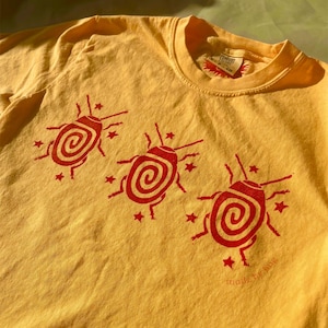 May include: A yellow short-sleeve t-shirt featuring a red graphic print of three insects with spiral patterns on their backs, surrounded by small stars. The interior neck label displays the text Comfort Colors and the size CH.