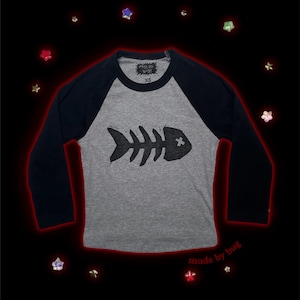 Puede incluir: Una camiseta de estilo raglán gris y azul marino con un diseño de espina de pescado negro. La camiseta de manga larga tiene el texto "made by bug" impreso en la parte inferior derecha. Talla XS.
