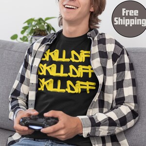 May include: Black t-shirt with the yellow text "SKILL OFF" repeated three times. The person is wearing a black and white plaid shirt over the t-shirt and is holding a black game controller. The background includes a gray couch and a plant.