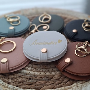 Könnte beinhalten: Sechs runde, kompakte Spiegel mit Schlüsselanhängern. Die Spiegel sind in verschiedenen Farben, darunter Blau, Weiß, Schwarz und Braun. Ein Spiegel trägt die Aufschrift "Brautmutter" in goldenen Buchstaben.