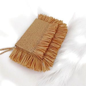 May include: A rectangular, woven raffia clutch in a natural tan color. The clutch features a fringe detail along the edges and a thin, woven strap. The bag is set against a white, fluffy background.