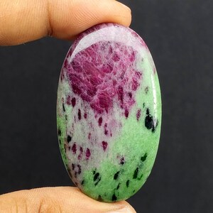 Puede incluir: Gema ovalada con una paleta de colores vibrantes. La piedra presenta una llamativa combinación de tonos rojo intenso y verde claro, con motas negras. La superficie pulida de la gema refleja la luz, resaltando su belleza natural. Una pieza única y llamativa.