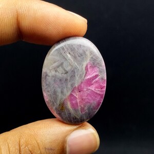 Peut inclure: Pierre précieuse ovale, mélangeant des teintes grises et roses vives. La surface polie révèle un motif naturel, avec le rose concentré dans une zone. La pierre est tenue entre les doigts sur fond sombre.