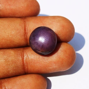 Op de afbeelding: Een dieppaarse ovale edelsteen met een glanzend, reflecterend oppervlak. De edelsteen wordt tussen twee vingers gehouden.