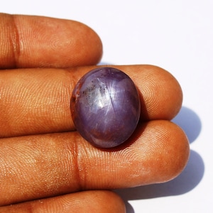 May include: A purple star sapphire gemstone with a star-like pattern in the centre. The gemstone is oval-shaped and has a smooth, polished surface.