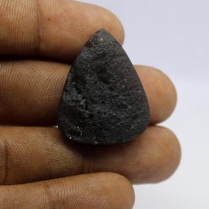 May include: A dark gray, teardrop-shaped gemstone with a rough, textured surface. The stone is held in a hand against a white background. The gemstone has a matte finish and appears to be a natural stone.