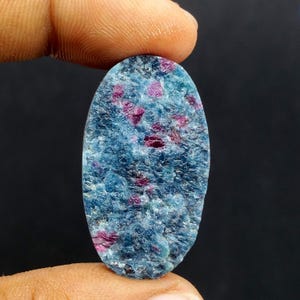 May include: An oval gemstone with a textured surface. The stone is mainly light blue, with small, irregularly shaped patches of deep red scattered throughout. The gemstone is held between two fingers against a dark background.