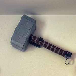 Op de afbeelding: Een display met Lego-modellen: Thor's hamer, Mjolnir, en twee Infinity Gauntlets. De hamer is grijs en bruin, de handschoenen zijn goud en rood. De items zijn op een witte muur gemonteerd, wat een superheldenthema cre&euml;ert.