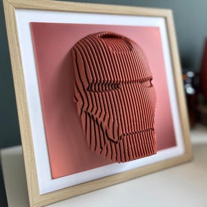 May include: A framed 3D wall art piece featuring a red, layered design of a superhero's face. The artwork is set against a red background and framed in a light wood frame.
