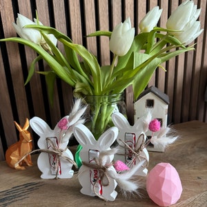 May include: Three white bunny-shaped ceramic holders with a brown and red chocolate bar inside each. The holders are decorated with a brown twine bow and a small bouquet of dried flowers. A pink geometric egg sits on a wooden table in front of the holders.