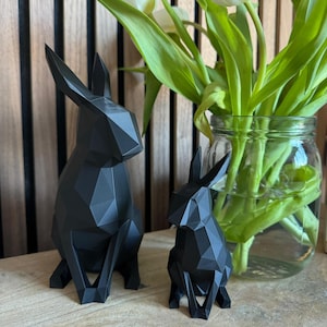 May include: Two black geometric rabbit sculptures, one larger than the other, sitting on a wooden surface. A vase of white tulips is behind the sculptures.
