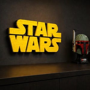 Pode incluir: Letreiro amarelo LEGO "STAR WARS" em uma parede preta. Tr&ecirc;s capacetes LEGO s&atilde;o exibidos em uma prateleira abaixo do letreiro. Os capacetes s&atilde;o Boba Fett, um Clone Trooper e um piloto imperial preto.