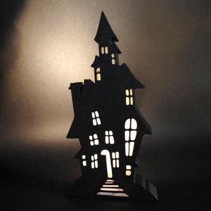 May include: A black wooden haunted house silhouette with cut-out windows and a lit interior. The house is standing on a black surface.