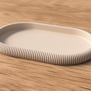 May include: A beige, oval-shaped tray with a ribbed edge. The tray is made of a solid material and sits on a wooden surface. The design is minimalist and modern, suitable for home decor or serving.