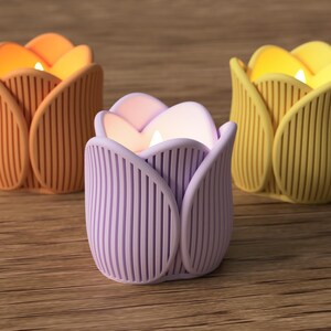 May include: Three tulip-shaped candle holders in orange, lavender, and yellow, each with a lit candle inside. The holders have vertical ribbed detailing and sit on a wooden surface. The candles emit a warm, inviting glow.