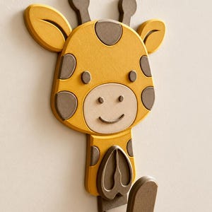 May include: A wall-mounted hook shaped like a cartoon giraffe head and neck. The giraffe is yellow with brown spots, brown ossicones, and a light beige muzzle. A brown hook extends from the bottom, featuring a heart-shaped detail on the front.