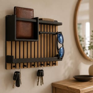 May include: A wall-mounted organizer featuring a light wood slatted back panel with a black metal frame. It includes a storage compartment, a shelf, and seven black hooks holding two sets of keys. A brown leather wallet and sunglasses are stored.