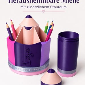 May include: A pencil-shaped desk organizer in shades of purple, pink, and light peach, featuring a removable top section, a central compartment for pens, and the name "Leya" embossed on the side. The text "Herausnehmbare Miene mit zus&auml;tzlichem Stauraum" is visible.