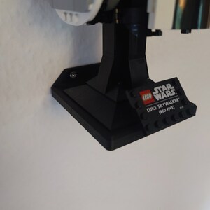 Puede incluir: Soporte de pared de pl&aacute;stico negro para un caza X-Wing LEGO Star Wars. El soporte tiene una base de ladrillo LEGO con el texto "LEGO STAR WARS LUKE SKYWALKER (RED FIVE)".