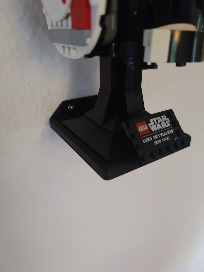 Puede incluir: Soporte de pared de pl&aacute;stico negro para un caza X-Wing LEGO Star Wars. El soporte tiene una base de ladrillo LEGO con el texto "LEGO STAR WARS LUKE SKYWALKER (RED FIVE)".