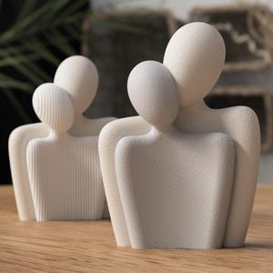 May include: Two off-white abstract sculptures of embracing figures. One has a textured surface, the other has vertical lines. The sculptures are on a wooden surface, with a blurred background of greenery and decorative items.