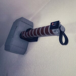 Op de afbeelding: Een grijze en bruine LEGO Mjolnir-hamer, gemonteerd aan een witte muur. De hamerkop is grijs met klinknageldetails en de steel is bruin met witte strepen. De steel heeft een zwarte band en het LEGO Infinity-logo.